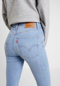 Jeans skinny de color azul claro con un acabado texturizado, que presentan un parche de cuero y la icónica etiqueta roja en los bolsillos traseros, mostrado en vista lateral.