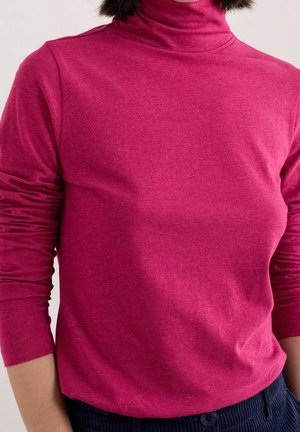 Osoba ubrana w różowy golf z długim rękawem i ciemnoniebieskie spodnie, widoczna od ramion do bioder.