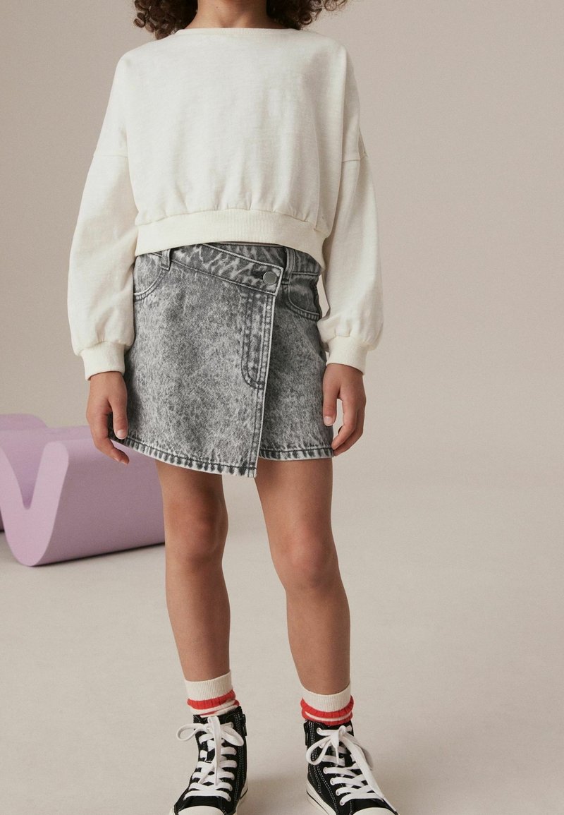 Enfant portant un haut à manches longues blanc, une jupe portefeuille grise effet délavé, des chaussettes rayées et des baskets montantes noires, debout à l'intérieur.