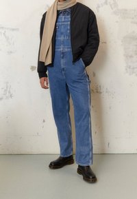 Černá bomber bunda přes modré denimové overaly s boční kapsou, hnědý šátek uvázaný kolem krku a černé chunky boty na podpatku.