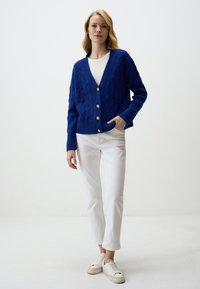 Cardigan blu in maglia con motivo a rombi, dotato di scollatura a V, cinque bottoni dorati, polsini a coste e vestibilità rilassata. Indossato sopra una maglietta bianca.