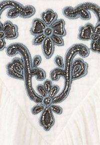 Tissu blanc avec une broderie florale grise complexe agrémentée de perles argentées. Le motif comprend des volutes et des formes de fleurs distinctes.