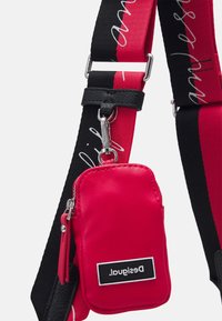 Punainen keinonahkapussukka, joka on kiinnitetty mustaan ja punaisiin säädettävään hihnaan. Siinä on hopeiset detaljit ja "Desigual"-merkintä. Pussukassa on teksturoituja yksityiskohtia.