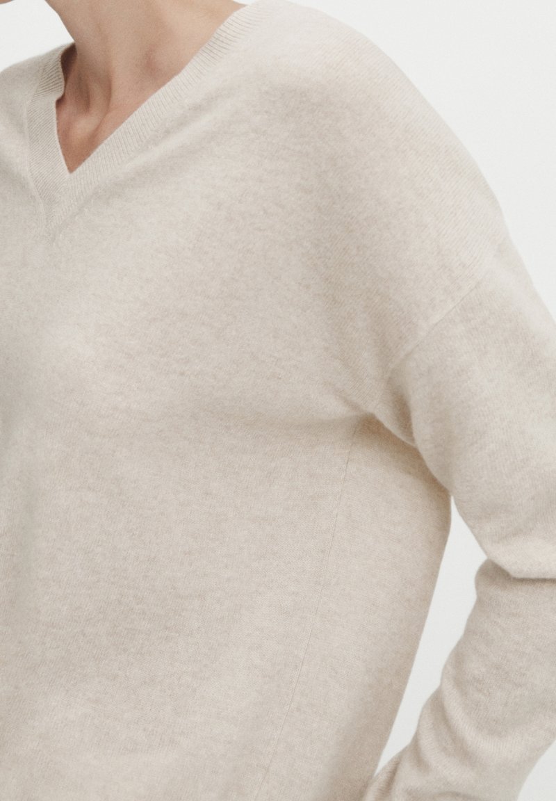 Personne portant un pull beige doux à col en V avec des manches longues sur un fond clair uni.