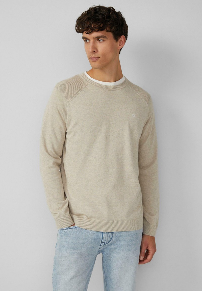 Beige Strickpullover mit Rundhalsausschnitt, gerippten Akzenten an den Schultern und langen Ärmeln. Getragen mit hellblauen Jeans. Dezentes Logo-Detail auf der Brust.