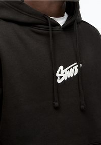 Černá mikina s kapucí vyrobená z měkké bavlny, která má bílé vyšívané logo. Obsahuje stahovací šňůrku na kapuci a má volný střih.