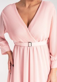 Różowa sukienka z głębokim dekoltem w kształcie V i plisowaną fakturą. Posiada długie rękawy oraz dopasowany pasek z kwadratową klamrą, podkreślający zwiewną spódnicę.