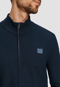 Jersey azul marino con cremallera y cuello alto, que presenta un parche de logo azul en el pecho y un diseño de tela texturizada.