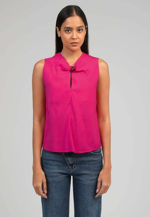 Femme portant un haut sans manches rose vif avec un fermoir décoratif au niveau du col et un jean bleu foncé, debout devant un fond gris uni.