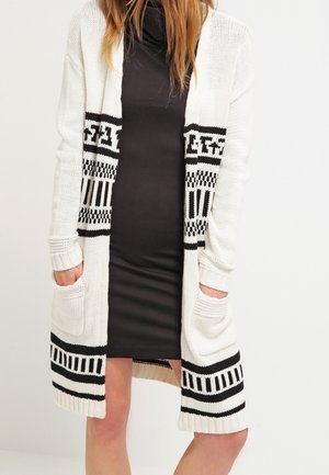 Frau trägt ein eng anliegendes schwarzes Kleid unter einer langen weißen Strickjacke mit schwarzen geometrischen Mustern und Taschen vorne.