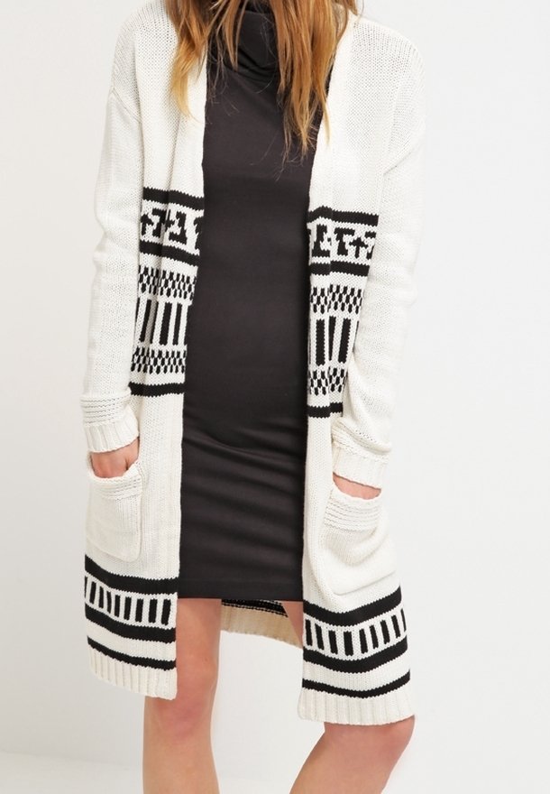 Femme portant une robe noire ajustée sous un long cardigan en maille blanche avec des motifs géométriques noirs et des poches avant.