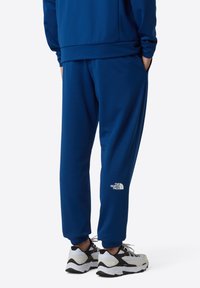 Blauwe joggingbroek met een elastische tailleband, zakken en een wit logo op het linkerbeen, gecombineerd met zwart-witte sportschoenen.