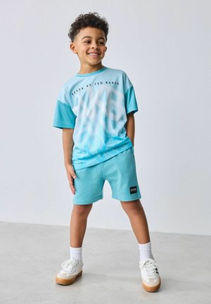 Smilende dreng med krøllet hår iført lys blå Ted Baker skjorte og shorts, stående med hænderne i lommerne mod en ensfarvet baggrund.