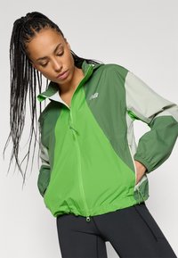 Chaqueta cortavientos verde y verde claro con cremallera frontal, puños elásticos y tejido ligero, que presenta un logo en la parte superior izquierda del pecho.
