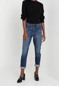 Dżinsowe spodnie w ciemnoniebieskim kolorze, proste nogawki, z lekko podwiniętymi końcami i delikatnym przetarciem; zestawione z czarnym swetrem i błyszczącymi czarnymi loafersami.