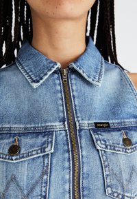 Gilet in denim di colore azzurro chiaro con colletto, zip frontale e due tasche sul petto. Presenta accessori in bronzo e un piccolo etichetta nera con la scritta "Wrangler".