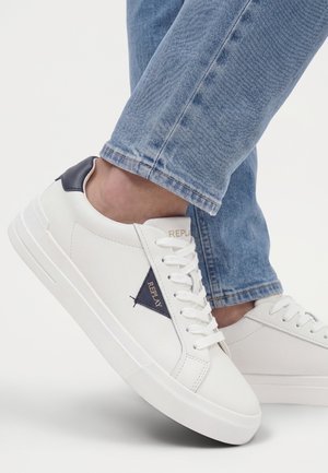 Baskets blanches avec logo bleu marine et détail au talon portées avec un jean bleu, jambes croisées sur un fond clair uni.