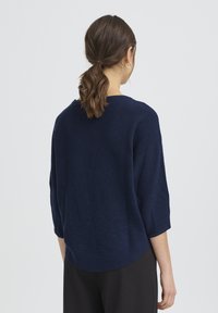 Pull en maille bleu marine avec manches trois-quarts, texture côtelée et ourlet arrondi, associé à un pantalon noir.