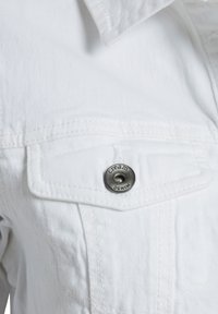 Bijeli traper jakna izbliza s prsnom džepom i srebrnim dugmetom s natpisom "Cream Denim", prikazujući teksturiranu tkaninu i detalje šivanja.