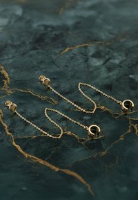 Guldkedjeörhängen med cirkulära accenter, med delikata länkar och transparent baksida på en mörkgrön marmoryta med guldådror.