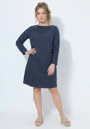 Žena s kudrnatými vlasy ve vysokém culíku, která má na sobě tmavě modré pruhované dlouhé šaty ke kolenům a béžové platformové sandály.