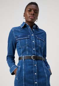 Jeansklänning i mörkblått, med knäppning fram, två bröstfickor, långa ärmar och ett svart bälte i midjan.