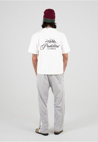 Hvit t-skjorte med svart tekst "Forbudt" og bladdesign på baksiden. Grå bukser med hvite sidestriper. Brune sko. Enkel, avslappet passform.