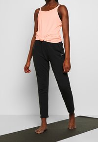 Top de melocotón con nudos en la cintura, combinado con pantalones jogger negros. Los joggers presentan un pequeño logo en el muslo izquierdo.