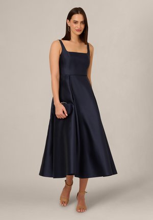 Kvinde iført ærmeløs navy blå satin midi kjole, holder matchende clutch og brune ankelremssko med hæl, poserer mod en ensfarvet baggrund.