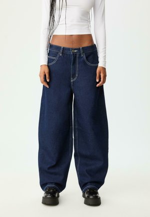 Jeans en denim bleu foncé à jambes larges avec des coutures contrastées, des poches avant et une fermeture à bouton, accompagnés de chaussures noires. Haut à manches longues blanc en dessus.