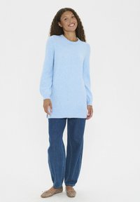 Kvinna stående, leende, iklädd en ljusblå långtröja, lösa mörkblåjeans och beige ballerinaskor med nittar.