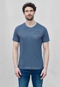 Blå bomull t-shirt med korta ärmar och v-ringning, med svart text som säger "LE SOLEIL TOUTE L'ANNEE" på den övre vänstra sidan.