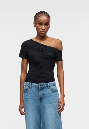 Femme aux cheveux tressés portant un haut noir asymétrique froncé et un jean taille haute bleu clair à jambes larges, debout devant un fond uni.