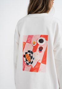 Witte oversized sweatshirt met een kleurrijke illustratie van voedsel op een geruit tafelkleed op de achterkant, met oranje, rood en zwarte accenten.