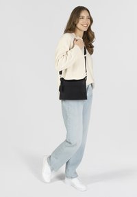 Zwarte leren schoudertas met verstelbare schouderband, subtiele logodetail en gladde textuur, gedragen met lichtblauwe jeans en een crèmekleurige trui.