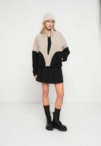 Μπεζ και μαύρο υπερμεγέθες μπουφάν με φερμουάρ μπροστά και υφή υφάσματος, συνδυασμένο με μαύρη φούστα και χοντρά μαύρα μποτάκια με καφέ κάλτσες.