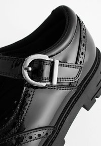 Chaussure de ville en cuir verni noir avec perforations décoratives, double surpiqûre et bride à boucle argentée sur semelle en caoutchouc noire.