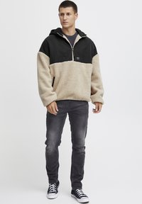 Felpa con cappuccio in pile, con la parte superiore nera e quella inferiore crema, con zip frontale e tasca frontale, abbinata a jeans grigi e sneakers nere con lacci bianchi.