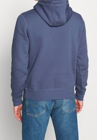 Felpa blu con cappuccio regolabile e polsini a costine, realizzata in materiale morbido. Mostrata indossata con jeans blu, vista posteriore che mette in evidenza cuciture e consistenza.