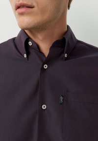 Dunkellila Hemd mit Knöpfen und Kragen, mit weißen Knöpfen und einem kleinen Logo auf der linken Brusttasche. Glatte Textur und maßgeschneiderte Passform.