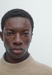 Zbliżenie osoby z krótkimi, teksturowanymi czarnymi włosami, ubraną w beżowy sweter z wysokim kołnierzem na ciemnej koszuli, na tle o neutralnym kolorze.