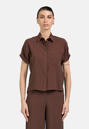 Donna con capelli tirati indietro che indossa orecchini a cerchio dorati e una camicia marrone a maniche corte abbottonata con pantaloni abbinati.