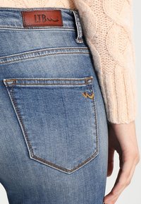 Blå denimjeans har en rundad bakficka med en brun läderetikett märkt "LTB" och subtila gula stickningsdetaljer.