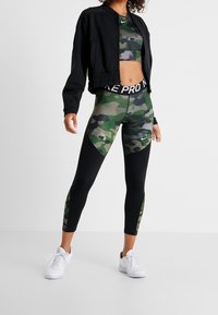 Conjunto de ropa deportiva que incluye una chaqueta bomber negra, un top corto de camuflaje verde y leggings a juego con acentos negros y cinturilla blanca con logotipo.
