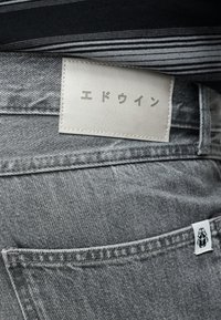 Κοντινό πλάνο γκρι τζιν τζιν με ραμμένη λευκή δερμάτινη ετικέτα που φέρει ιαπωνικό κείμενο και ένα μικρό λογότυπο σκαθαριού στην ετικέτα της πίσω τσέπης.