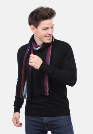Jeune homme en pull noir et jeans ajustant une écharpe à rayures multicolores, souriant et regardant sur le côté devant un fond blanc.