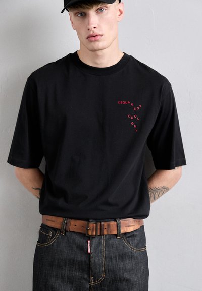 Ein junger Mann trägt eine schwarze Kappe, ein übergroßes schwarzes T-Shirt mit rotem Text, einen braunen Ledergürtel und dunkle Jeans vor einer schlichten grauen Wand.