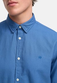 Camisa abotonada de color azul claro con cuello, presenta botones blancos y un pequeño logo bordado en el lado izquierdo del pecho. Textura suave.