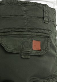 Olive vihreät cargo-shortsit, joissa on läppätasku ja nahkainen logo-label. Kangas on sileä, ja siinä on näkyvät sauman yksityiskohdat ja tekstuuri.