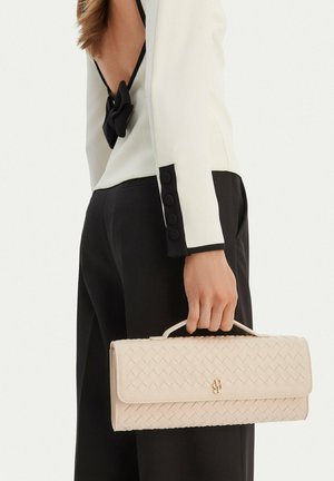 Kobieta w czarno-białym stroju trzyma kremową plecioną torebkę z uchwytem na górze, pokazując tylny, odsłonięty detal oraz mankiet z zapięciem na guziki.